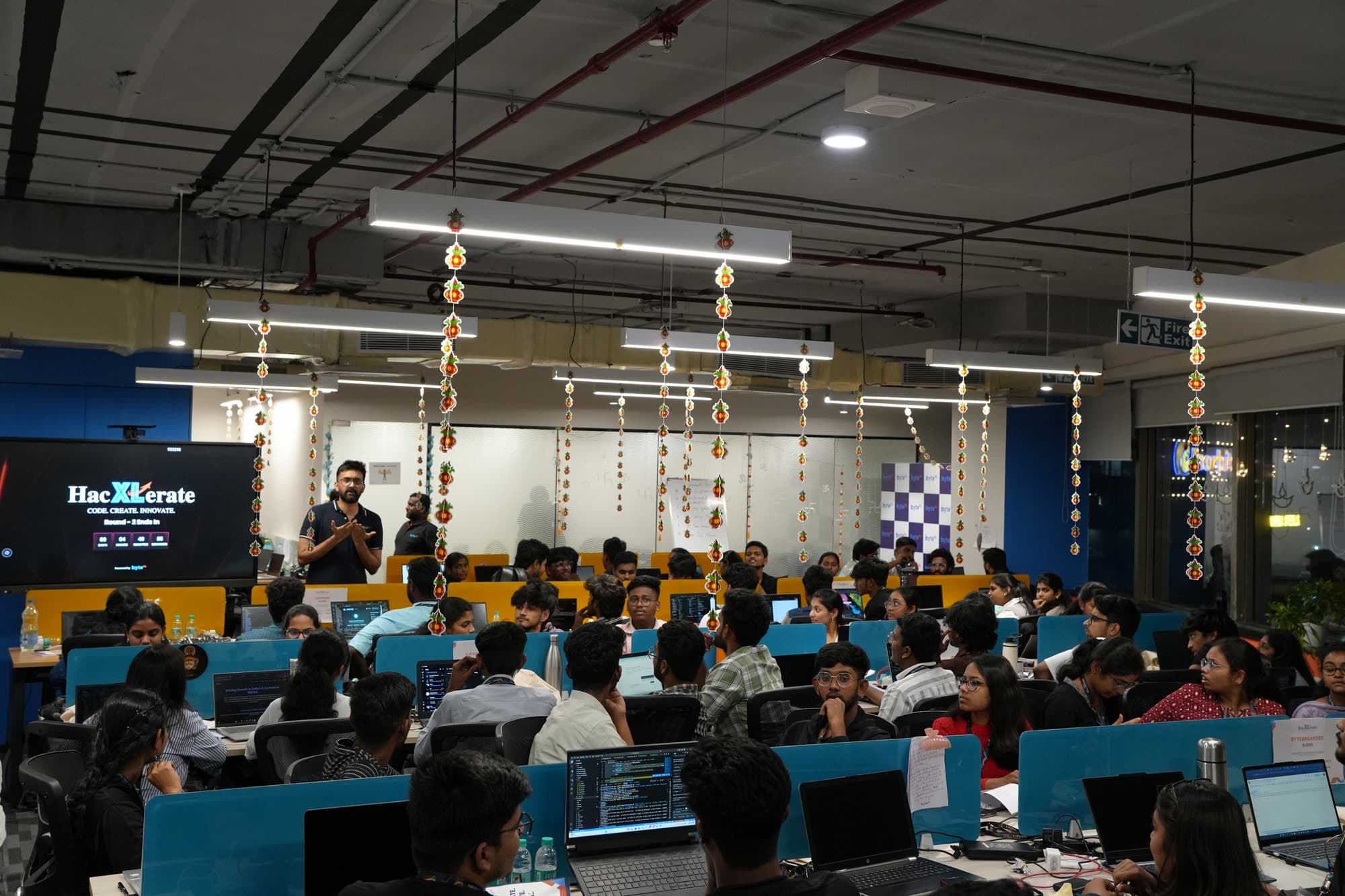 A packed HacXLerate coding floor with teams building at their desks under festive lights