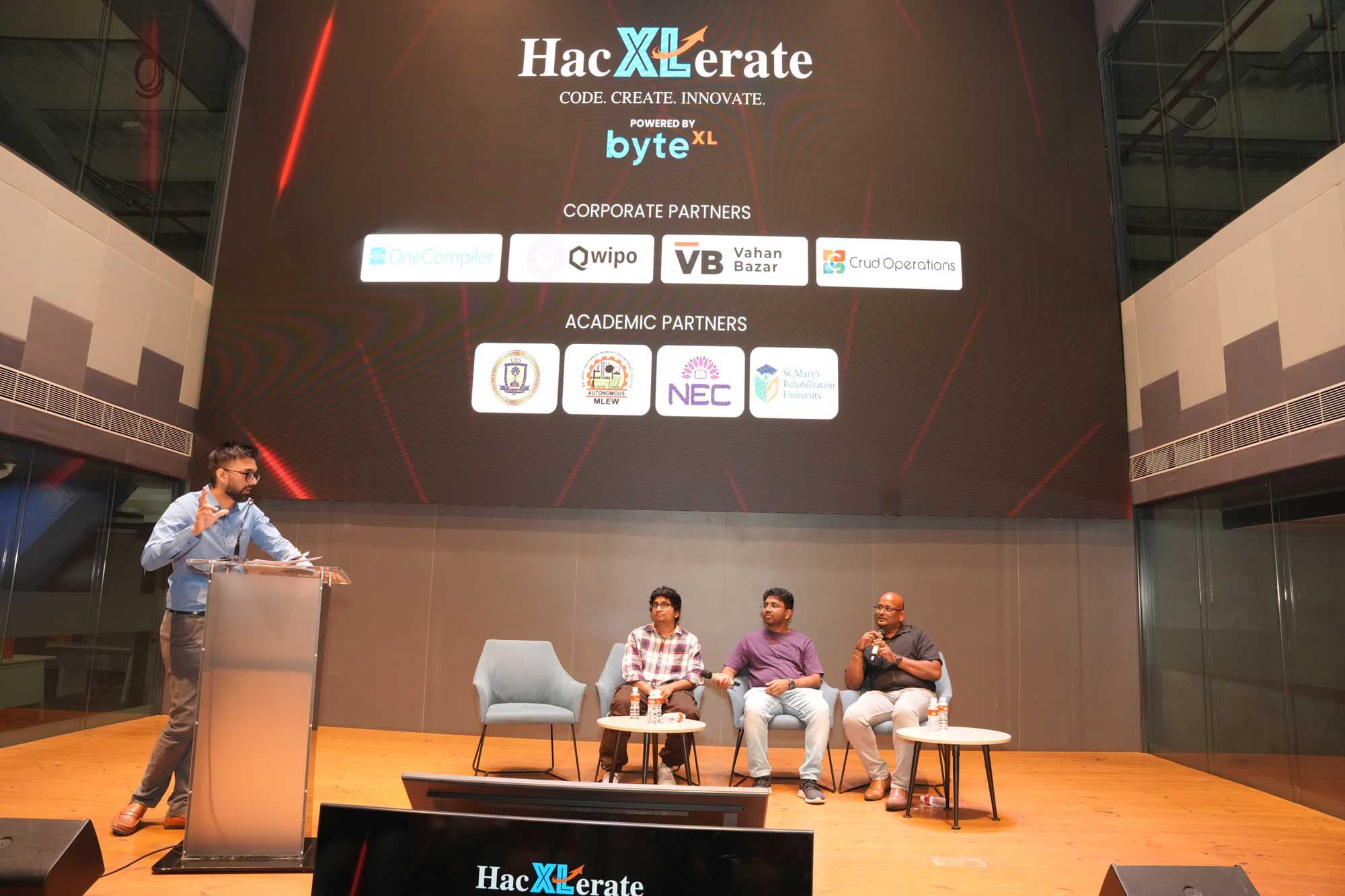 A speaker addressing the audience beneath the HacXLerate partner board