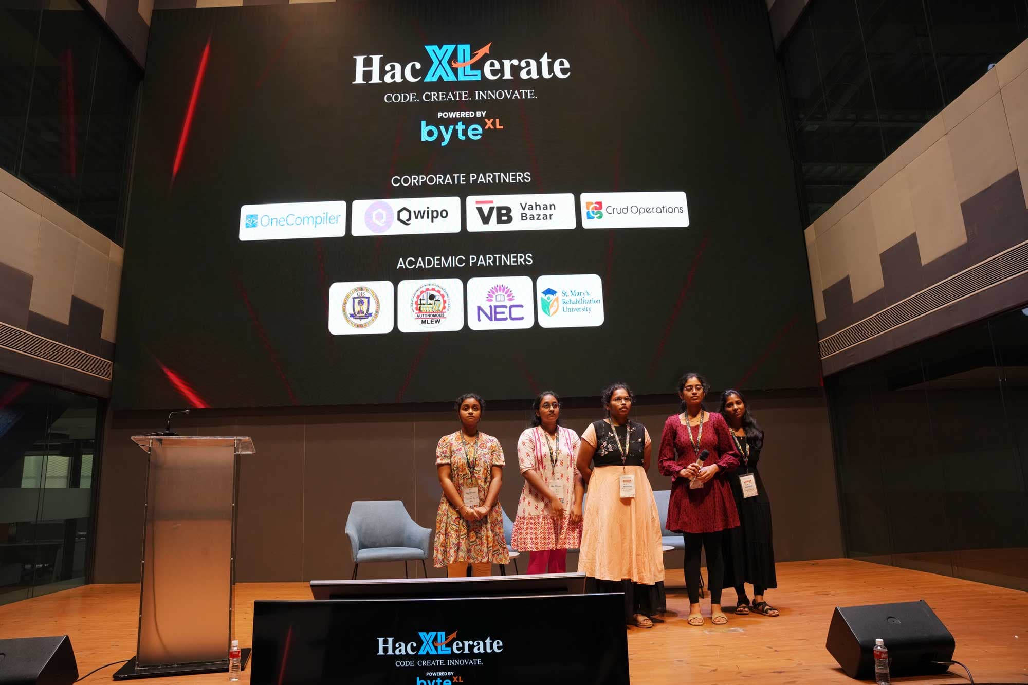 A team on stage in front of the HacXLerate partner board